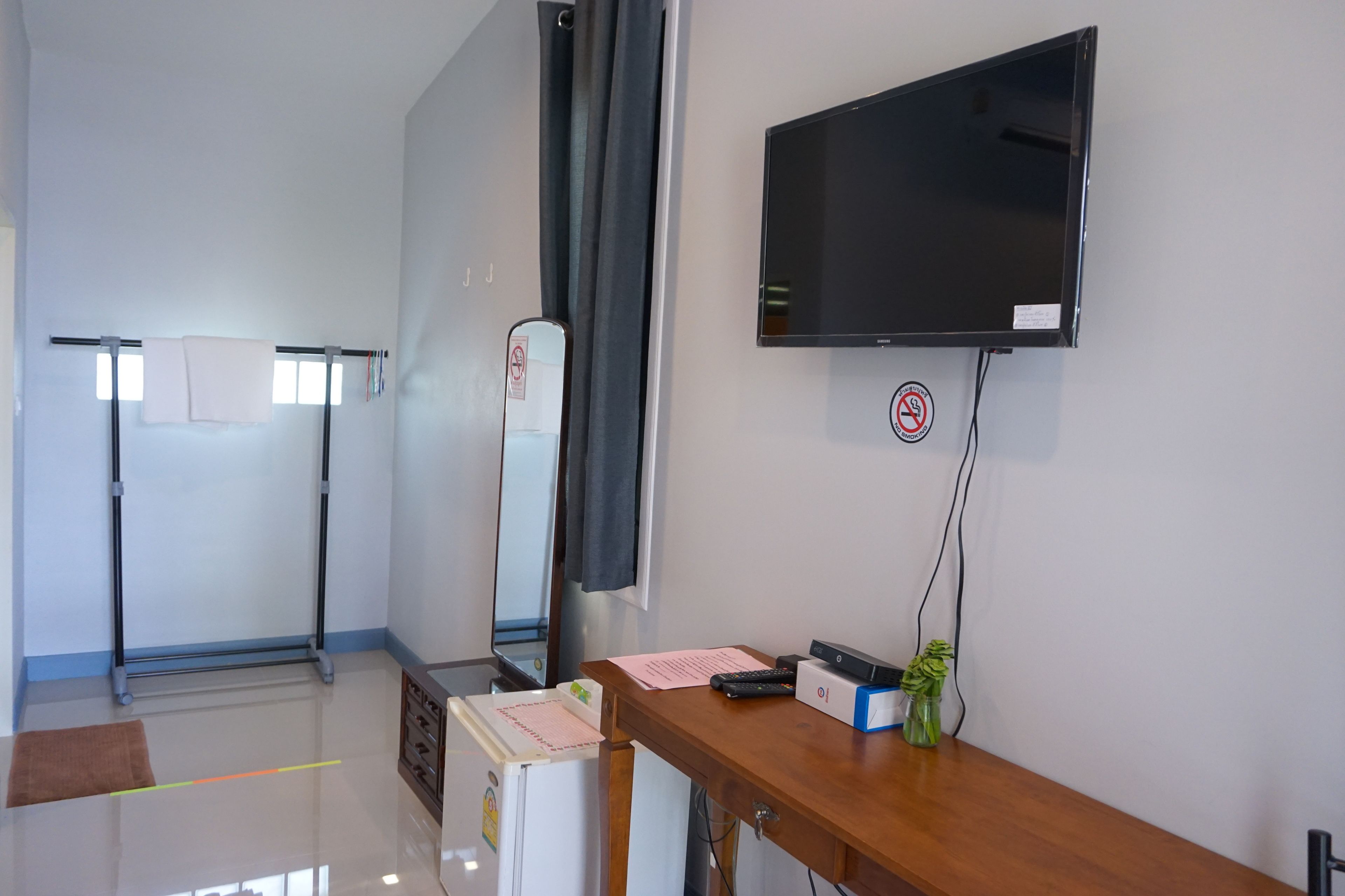 Quarto de hotel com televisão de tela plana na parede, escrivaninha de madeira com teclado, uma pequena planta e um espelho.