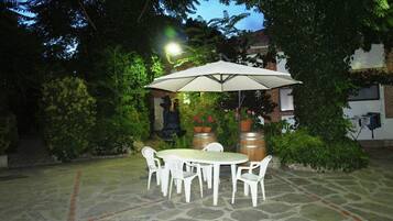 Un patio al aire libre con suelo de piedra, sillas de plástico blanco, una mesa redonda y una sombrilla grande que da sombra.