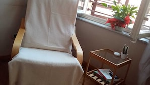 Una silla de madera con funda blanca, una mesita de madera con un libro y un cactus, y una ventana con una planta.