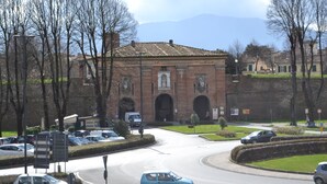 Una glorieta con un edificio histórico que presenta entradas arqueadas, rodeada de coches aparcados y árboles.