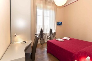 A hotel room with a bed, a desk, a chair, a TV, and a window with sheer curtains. - Nonna Adriana (Lucca)