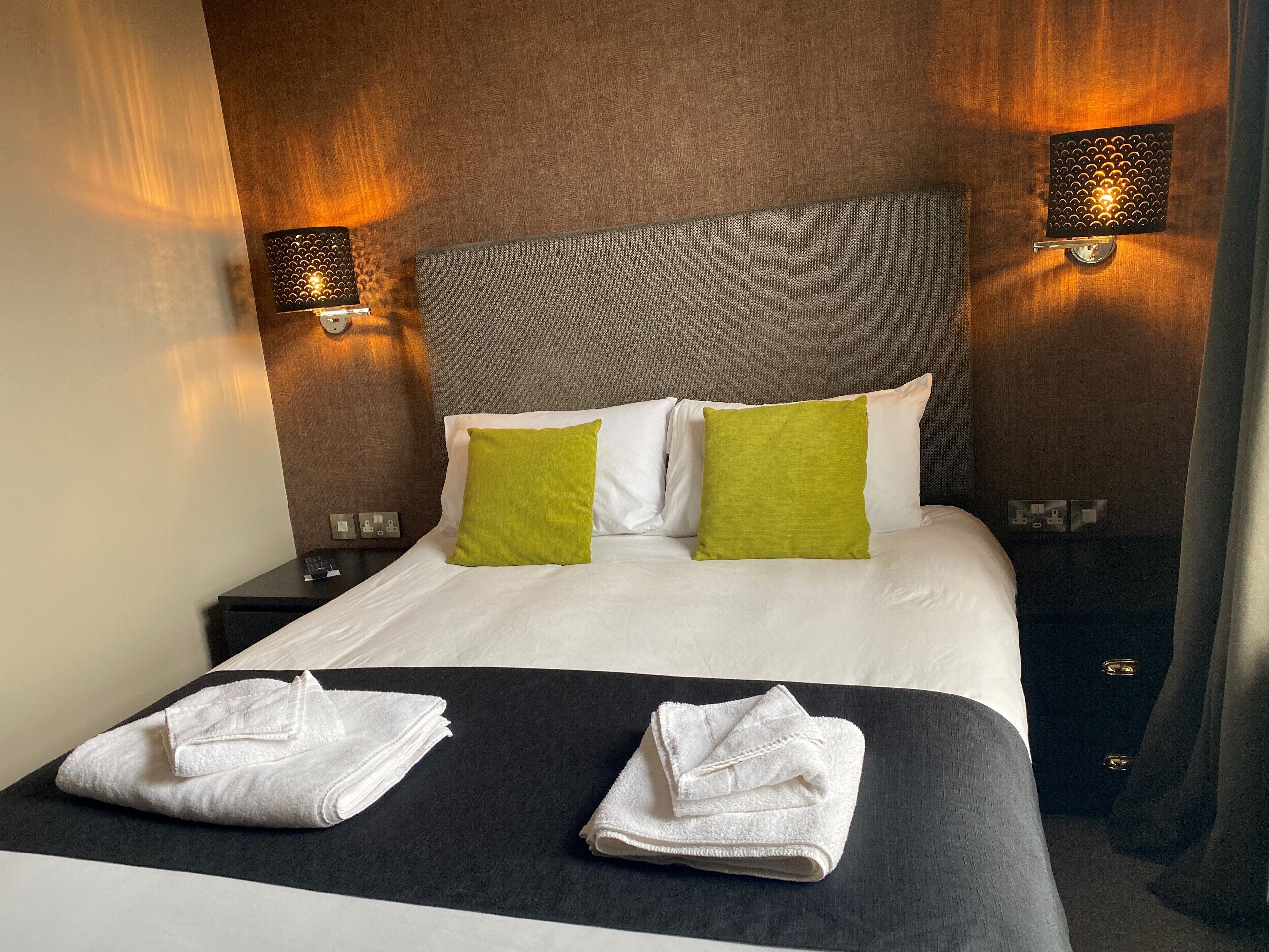 Une chambre d’hôtel avec un lit bien fait, deux oreillers, une tête de lit noire et deux lampes fixées au mur.
