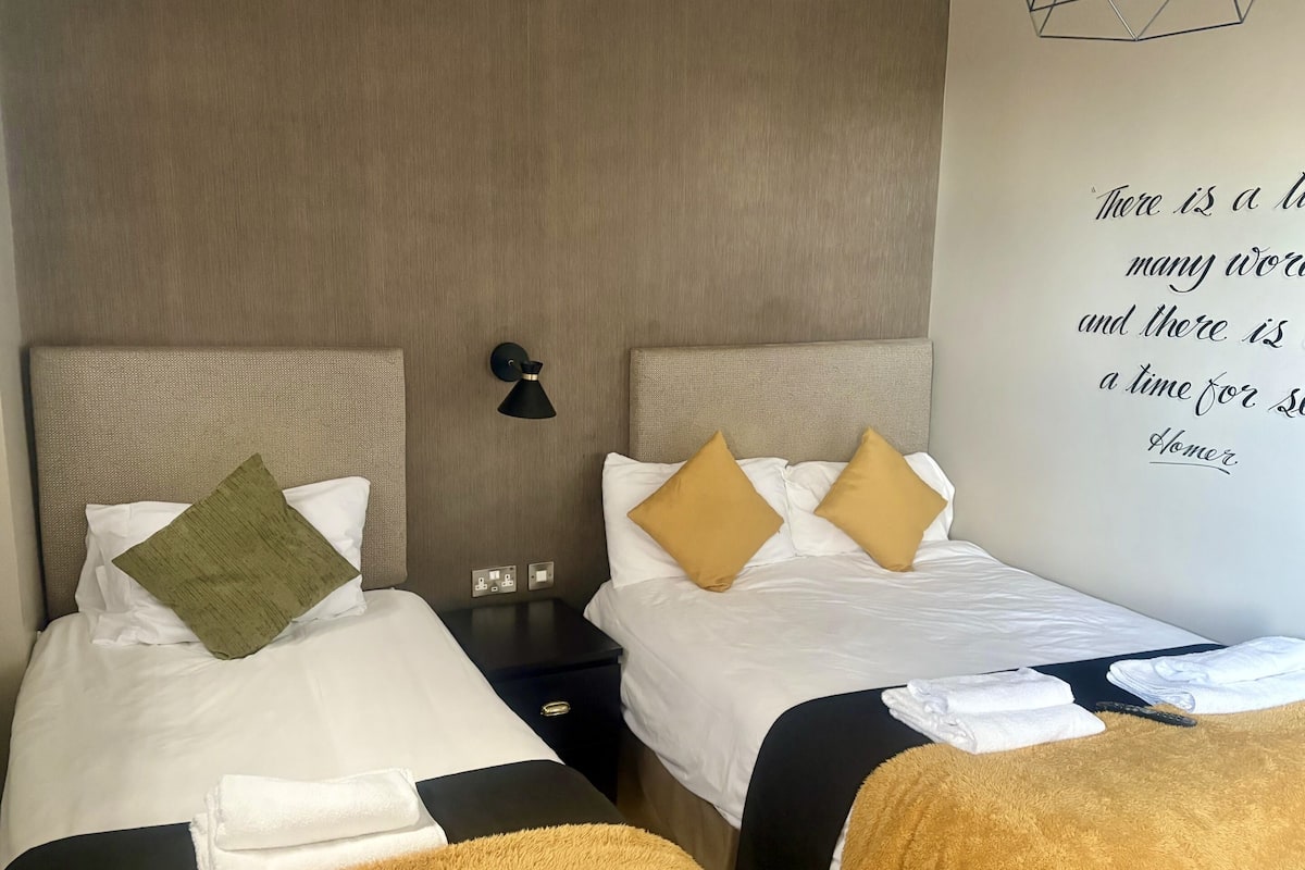 Quarto de hotel com duas camas, cada uma com roupa de cama branca e almofadas decorativas. Há uma luminária na parede entre as camas, e uma frase na parede.