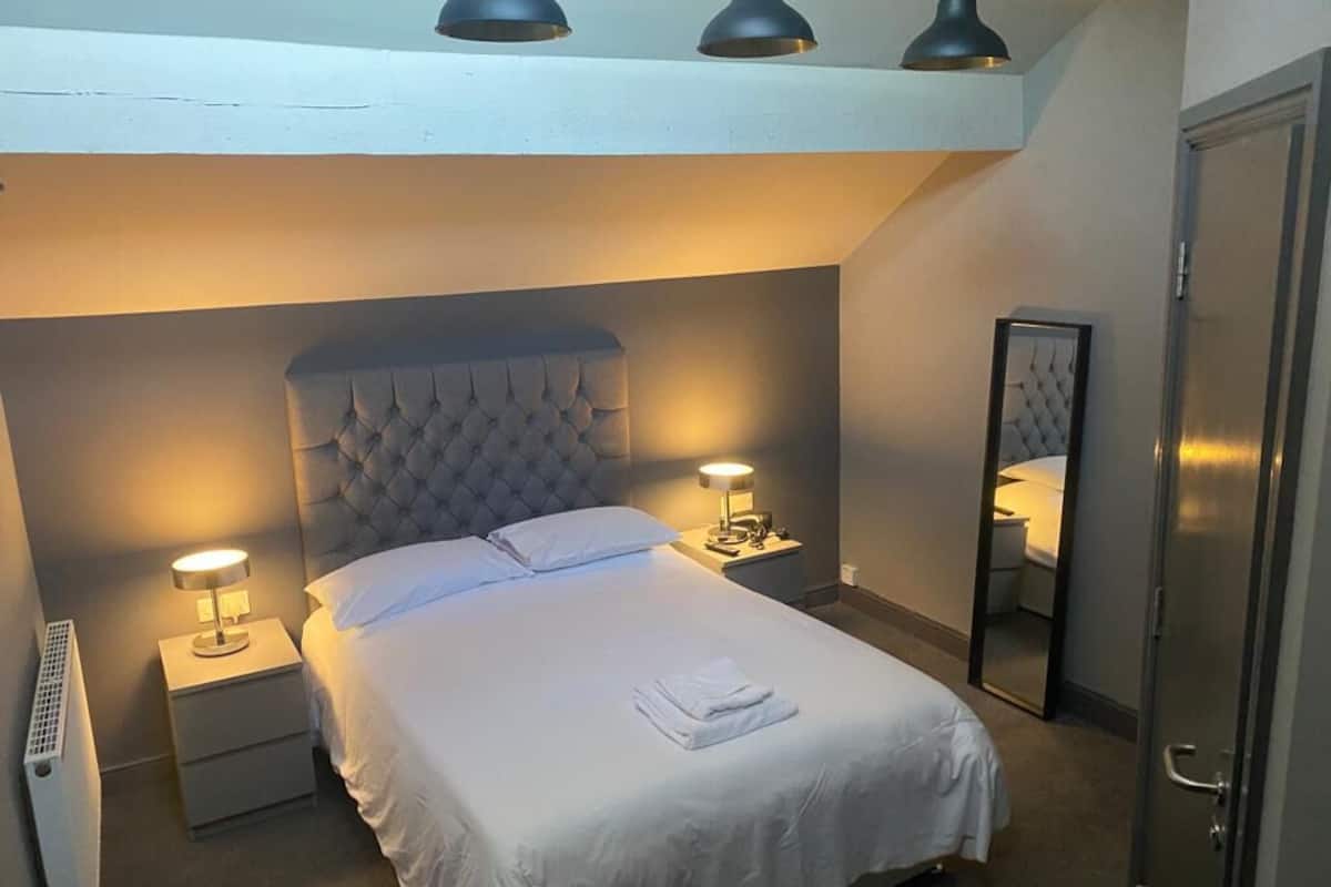 Uma cama bem arrumada com cabeceira estofada, duas luminárias de apoio e um espelho na parede.