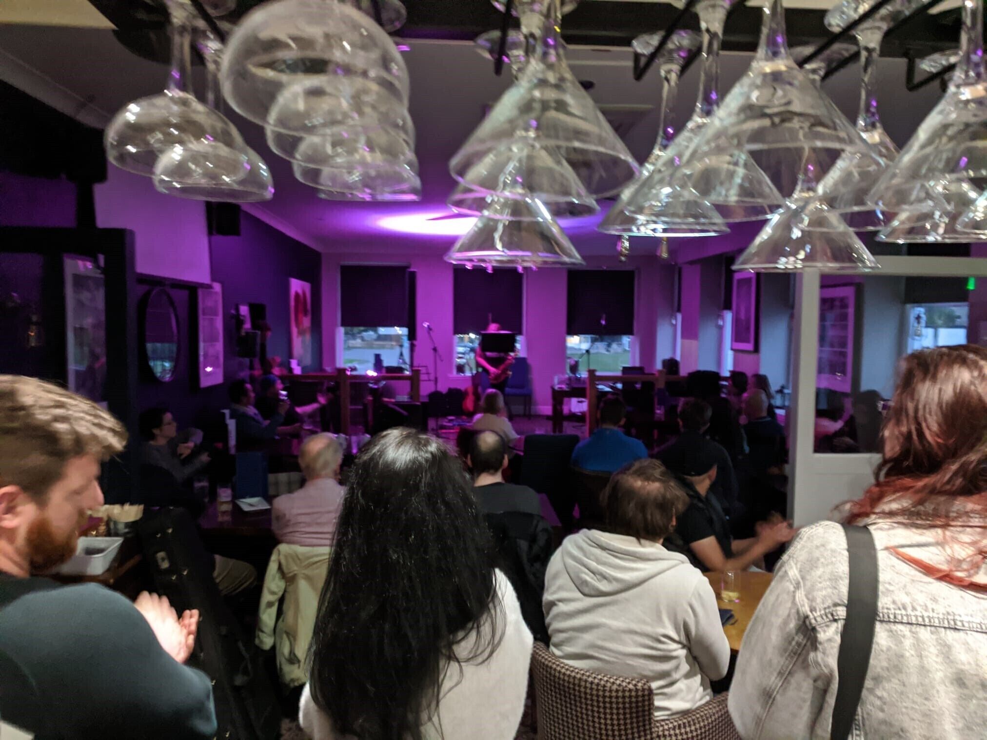 A crowded indoor space with hanging glass chandeliers, a stage with a performer, and seated patrons facing the stage.