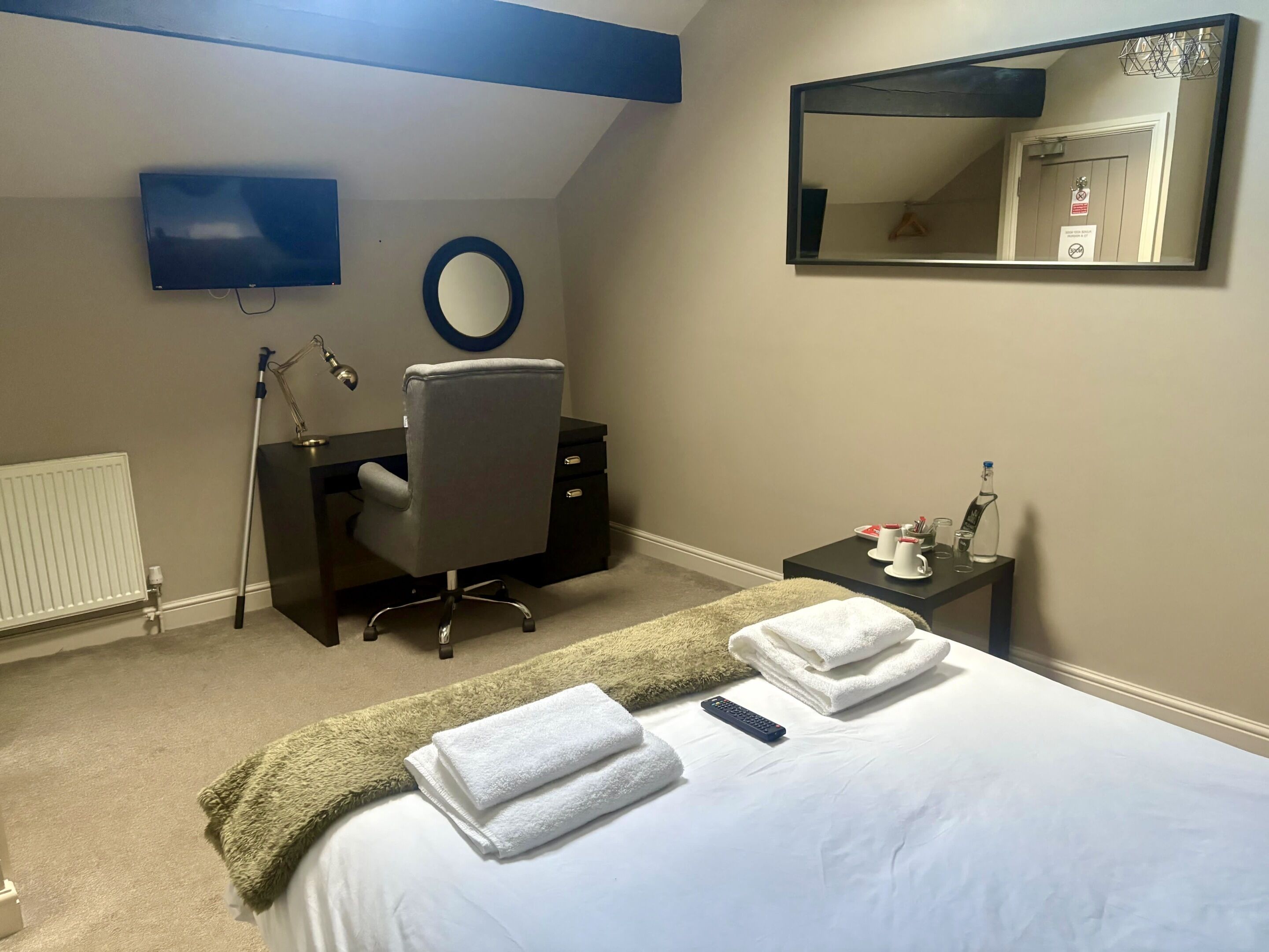 A hotel room with a bed, a desk, a chair, a TV, a mirror, a radiator, and a small table with toiletries.