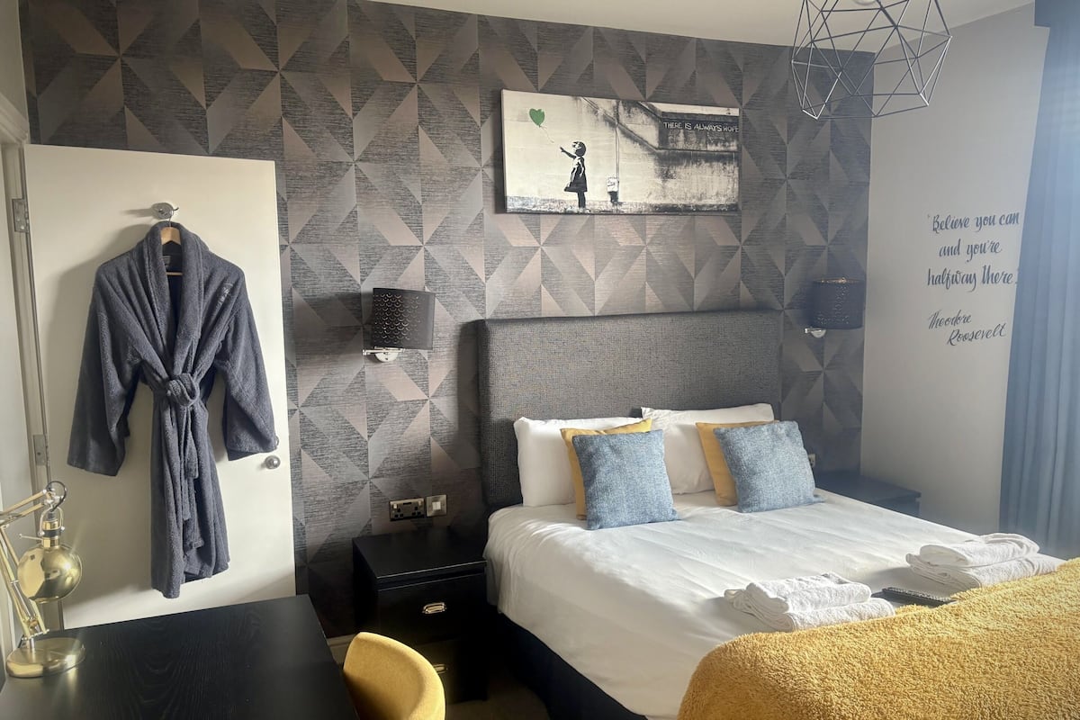 Quarto de hotel com parede estampada em padrão geométrico, cama com roupa de cama branca e azul, mesa de cabeceira com abajur, roupão pendurado na porta e uma frase motivacional na parede.