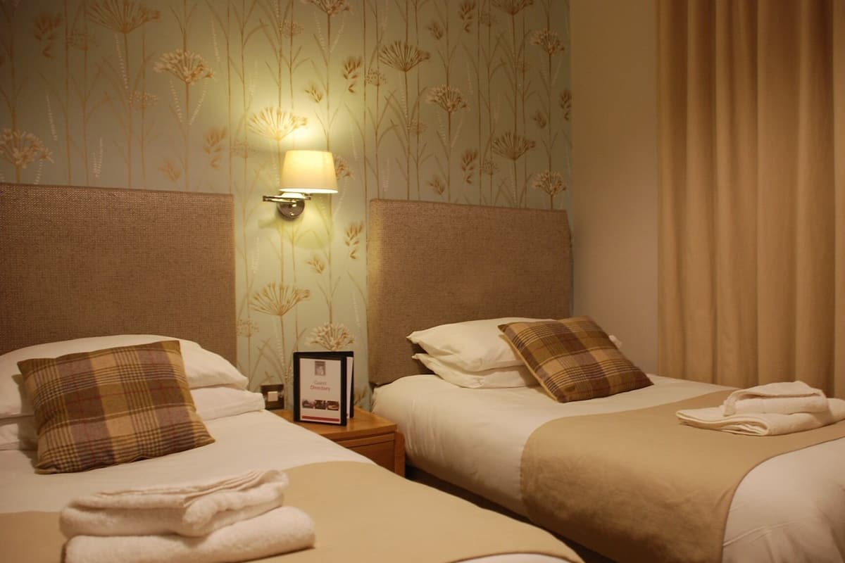 Quarto de hotel com duas camas, cada uma com um travesseiro, uma mesinha de cabeceira com abajur e um quadro emoldurado.