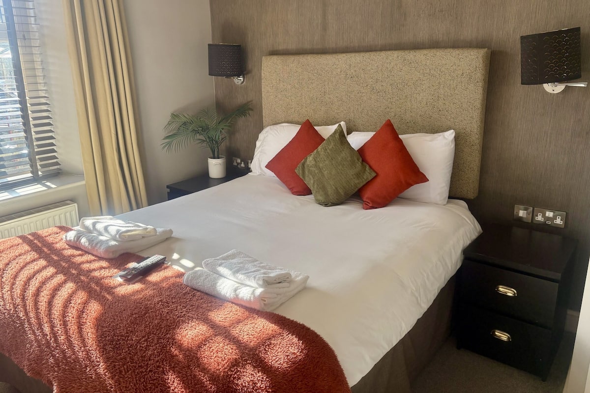 Cama bem arrumada com lençóis brancos, travesseiros vermelhos e verdes, e um cobertor vermelho. Há uma pequena planta em vaso sobre uma mesa de cabeceira, uma luminária na parede e uma tomada na parede.