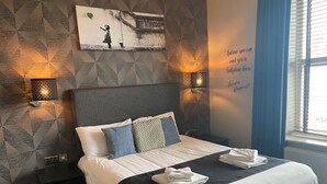 A hotel room with a bed, pillows, a wall mural, two wall sconces, a window with blinds, and a blue chair. - The White Swan (Leeds)