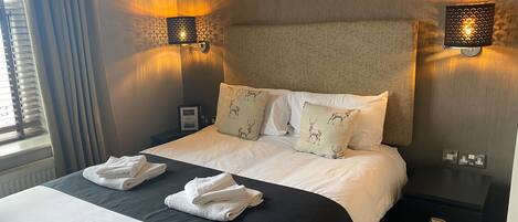 A neatly made bed with white linens and two decorative pillows, a bedside table with a lamp, and a power outlet on the wall.