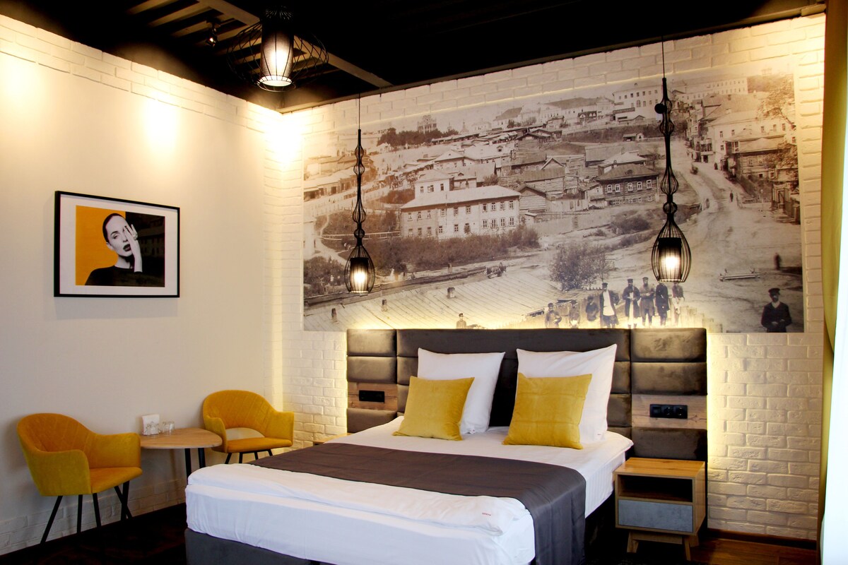 A hotel room with a large bed, two yellow chairs, a small table, and a cityscape mural on the wall.