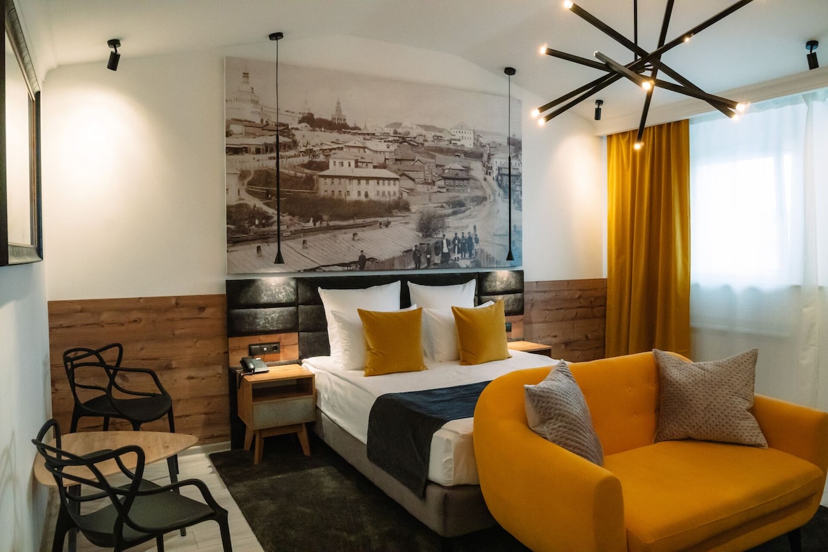 A hotel room with a bed, a yellow armchair, a small table with chairs, a nightstand, and a large cityscape painting on the wall.