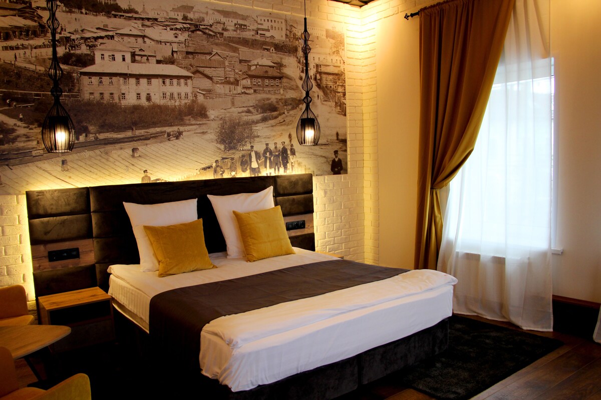 A hotel room with a large bed, a headboard with a cityscape mural, two bedside lamps, and a window with sheer curtains.