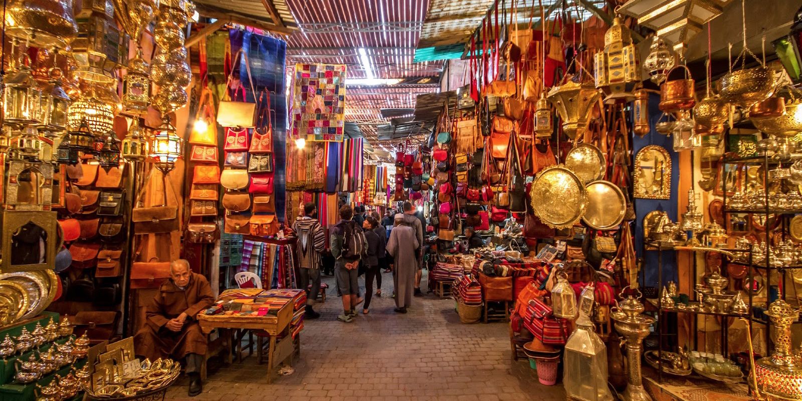 A bustling market with various stalls selling bags, textiles, and metalware.