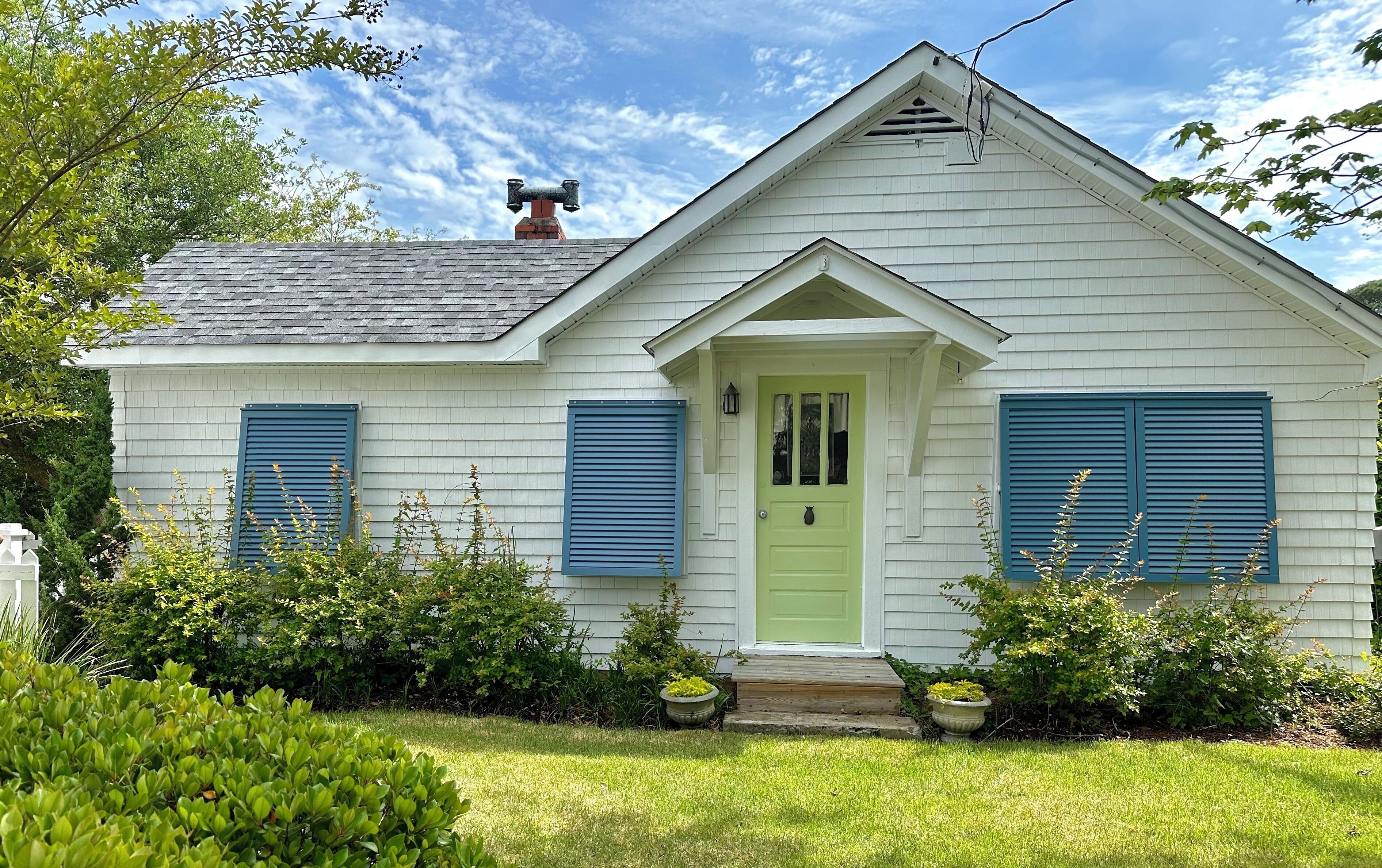 A white house with green shutters and a green door, surrounded by greenery and a clear sky.