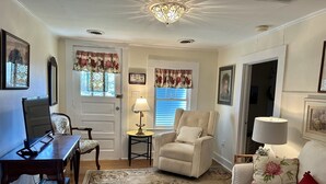 A living room with a sofa, armchair, and a small table with a lamp. There is a door with floral curtains and a window with blinds.