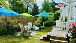 A backyard with a wooden deck, white outdoor furniture, and a small garden area with potted plants and flowers.