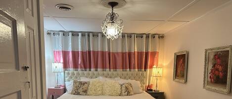 Un dormitorio con cama, mesita de noche, silla, candelabro y ventana con cortinas.