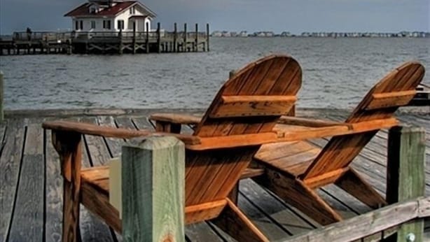 A wooden pier with three wooden chairs facing the water. In the background, there is a lighthouse on a pier.