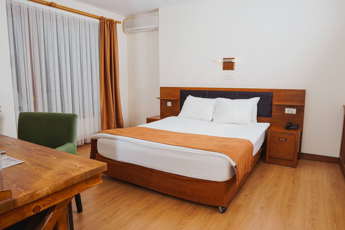A hotel room with a wooden bed, a green chair, a wooden desk, and a nightstand with a phone.