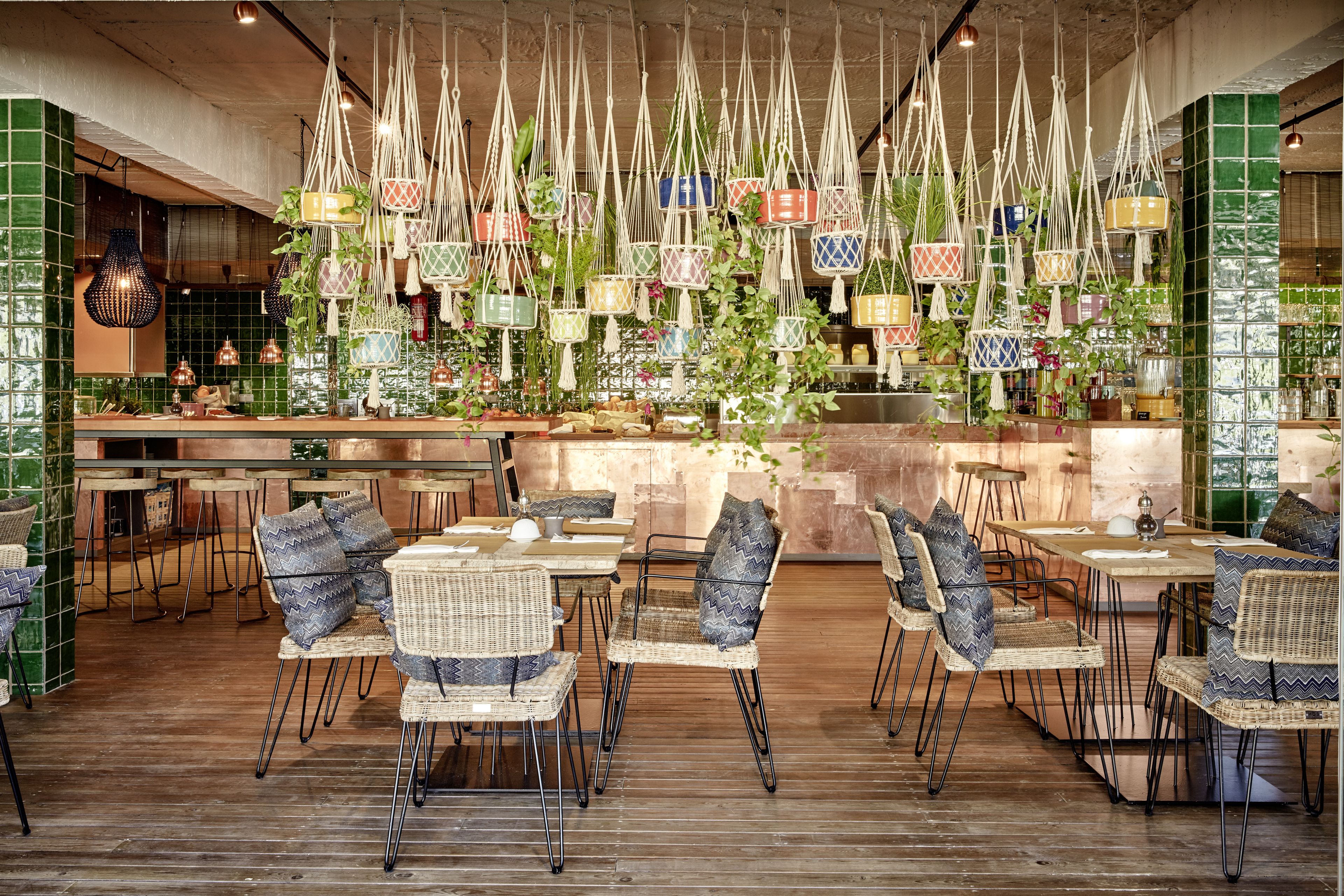 Un restaurante con sillas y mesas de mimbre, macramé colgante como decoración, y un salpicadero de azulejos verdes.