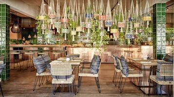Un restaurante con sillas y mesas de mimbre, macramé colgante como decoración, y un salpicadero de azulejos verdes.