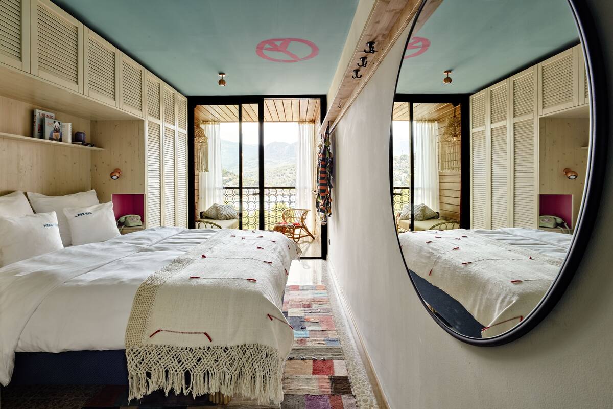 A bedroom with a large mirror, a bed with white linens, a colorful rug, and a view of the outdoors through a sliding door.