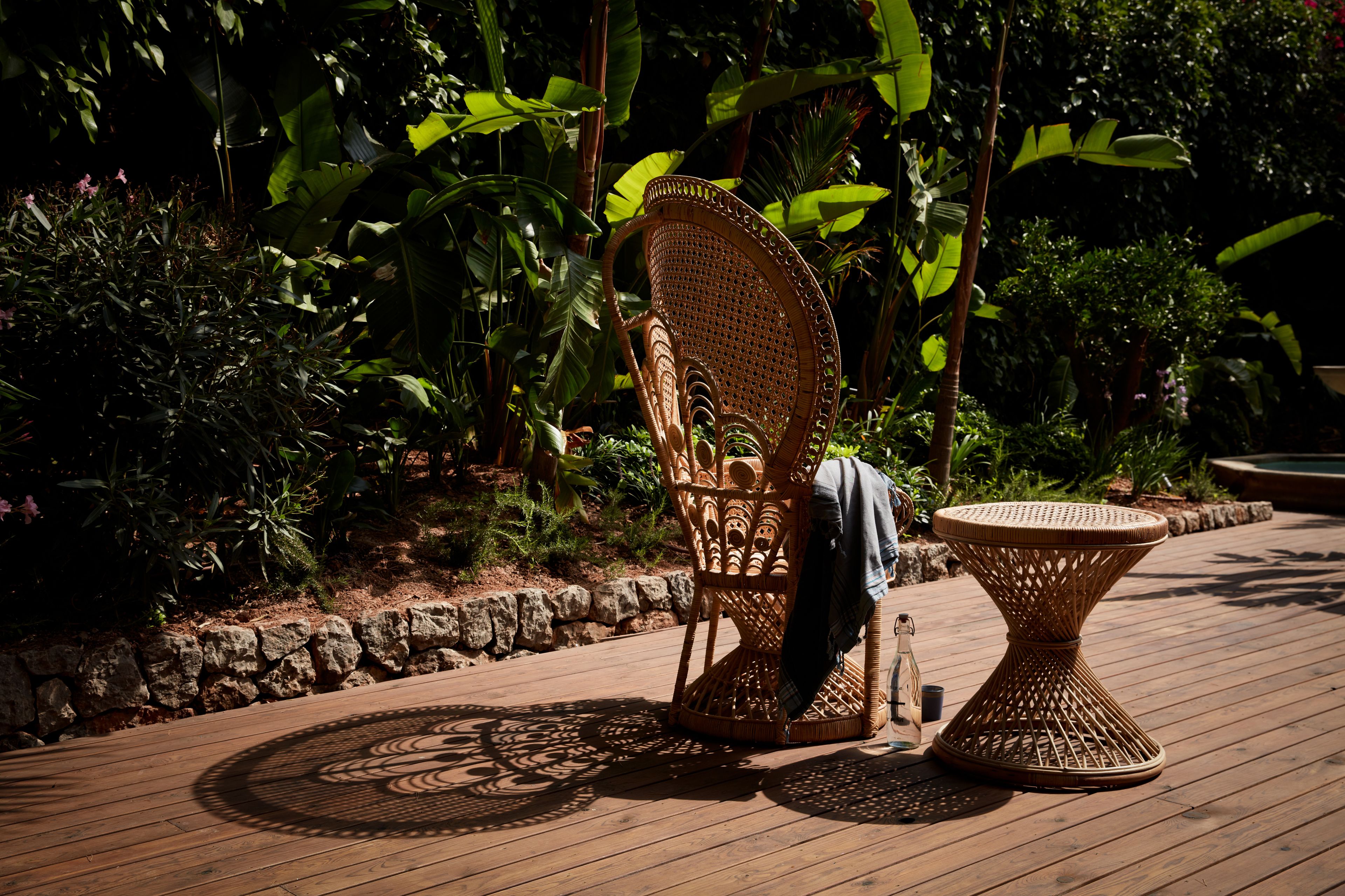 Een rotan stoel en tafel staan op een houten terras, omgeven door weelderig groen.