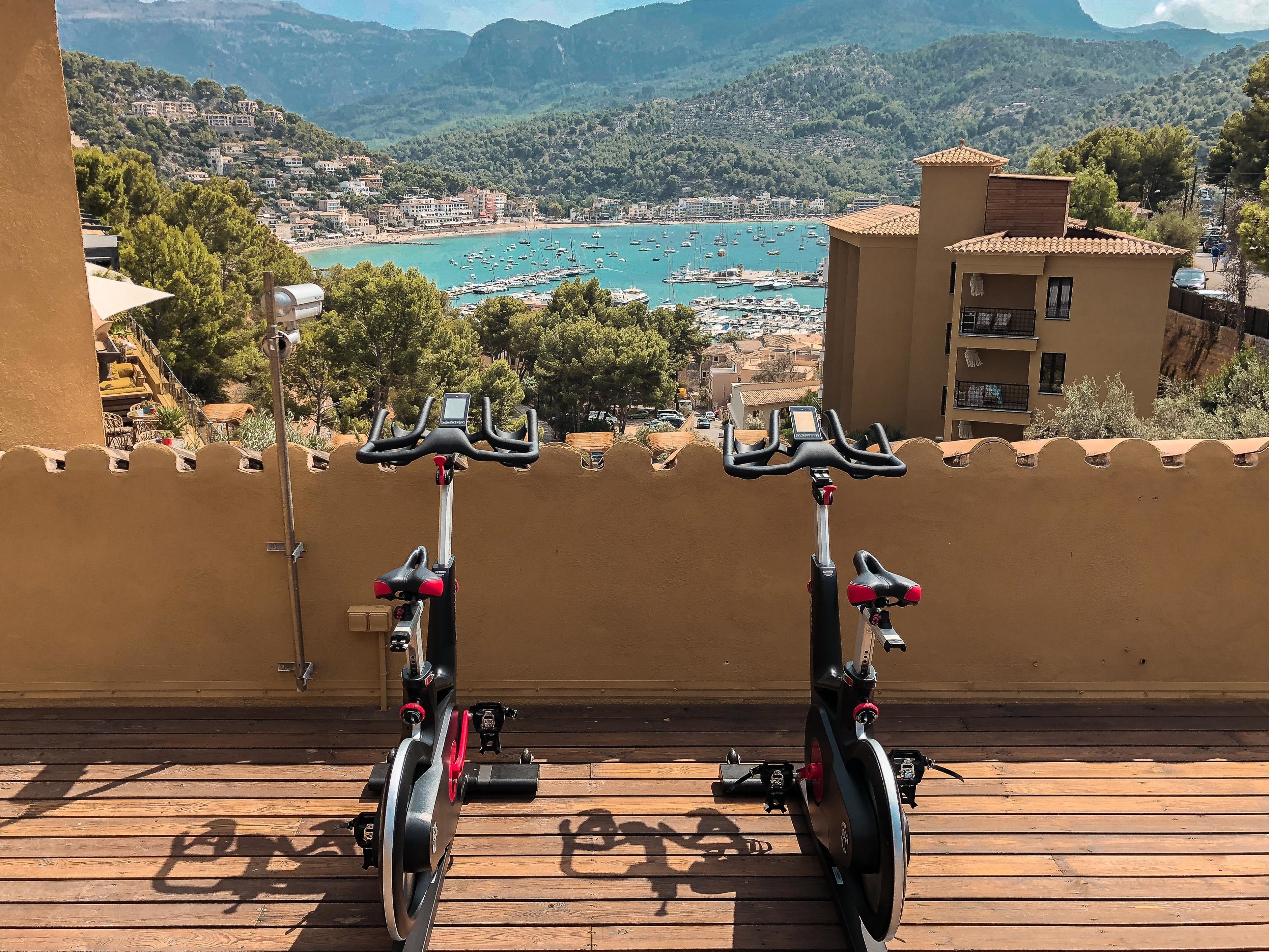 Deux vélos d’appartement sont installés sur une terrasse en bois, offrant une vue panoramique sur une ville côtière et des montagnes en arrière-plan.