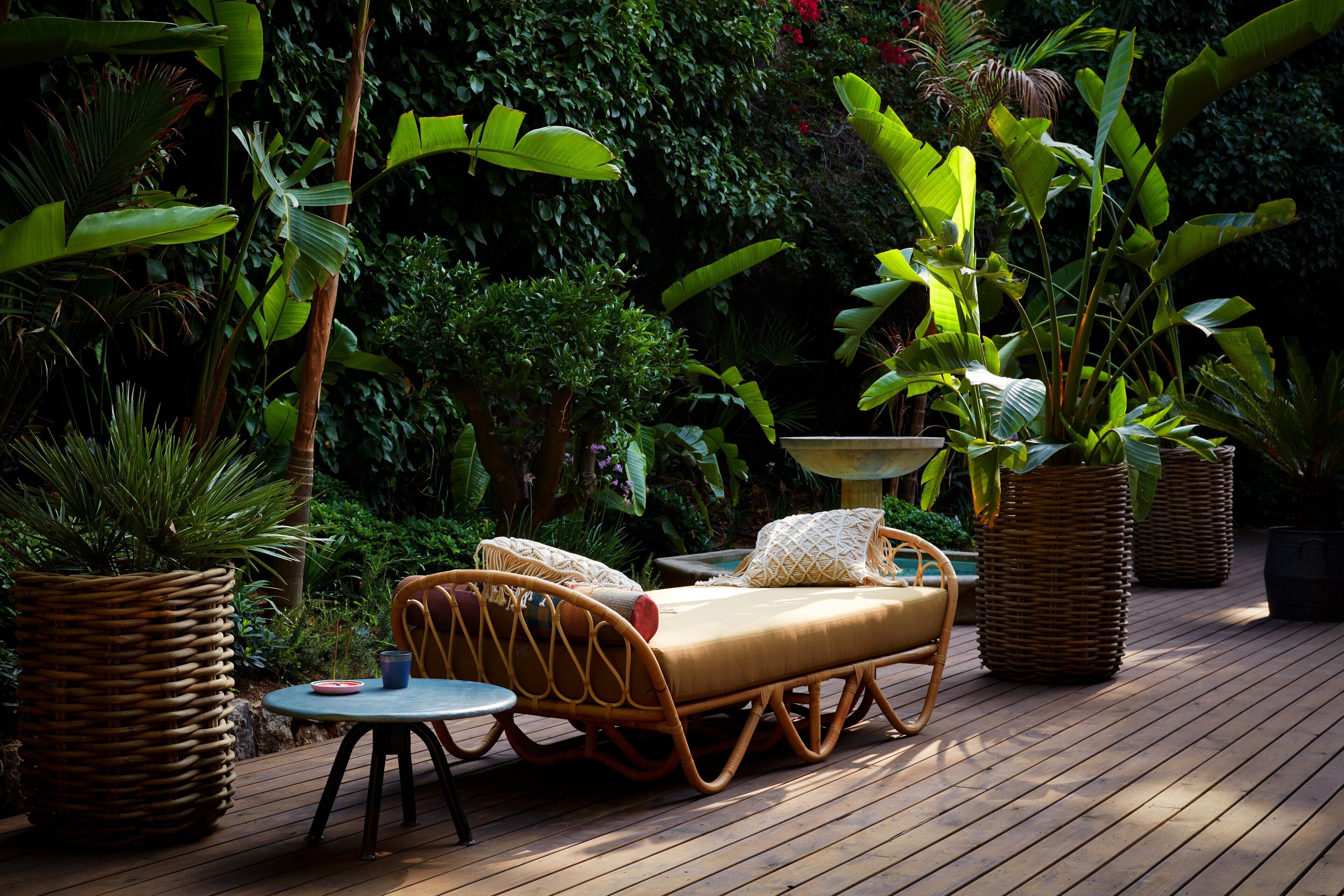 A wicker lounge chair with cushions, a small round table, and a potted plant in a tropical garden setting.