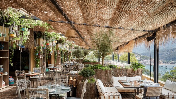 An outdoor dining area with wicker furniture, hanging planters, and a view of the city.