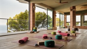 A spacious room with large windows offering a view of the sea and trees. There are yoga mats and colorful cushions on the floor.