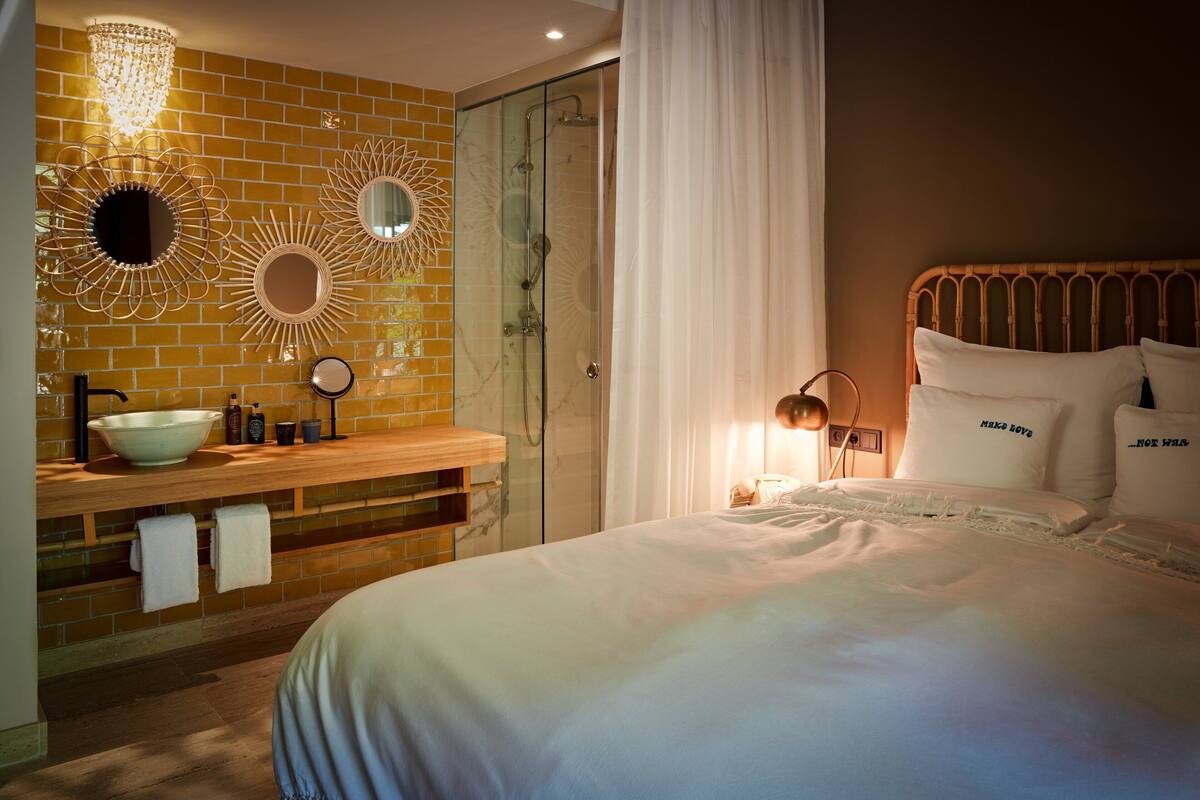 A hotel room with a large bed, a wooden vanity with a bowl sink, two wall mirrors, a chandelier, and a glass shower enclosure.