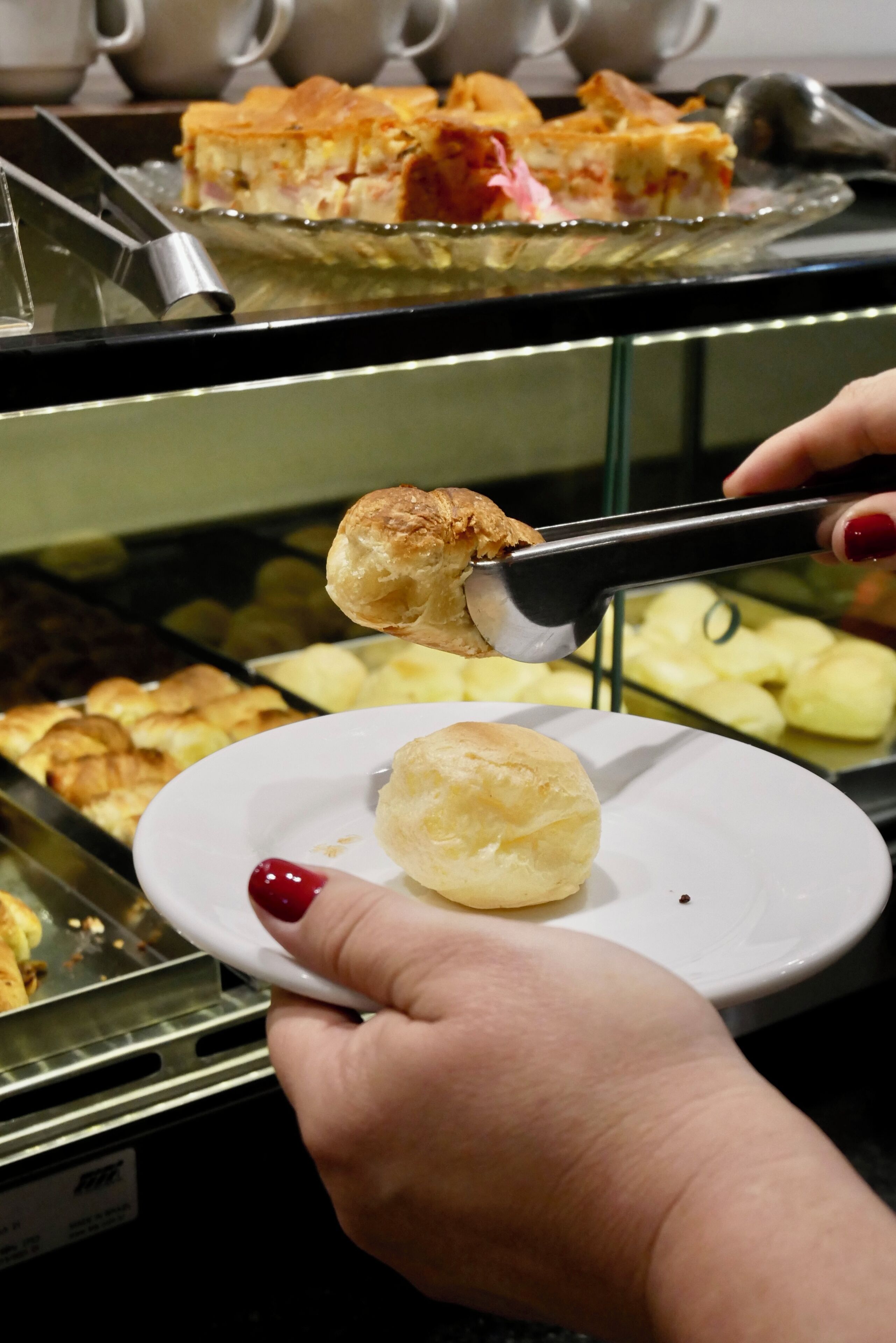 Una mano sosteniendo unas pinzas sobre un plato con un pastel, con una exhibición de productos horneados al fondo.