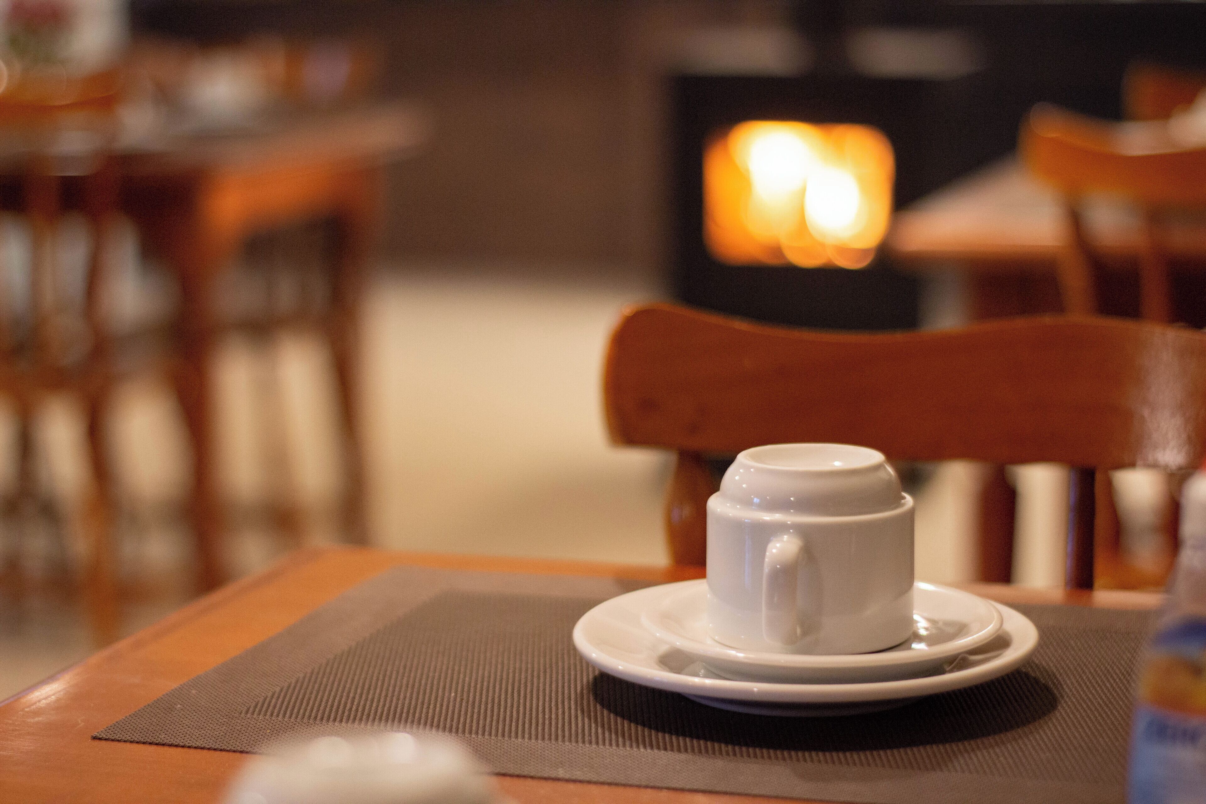 A white cup on a saucer on a table with a placemat, a wooden chair, and a blurred background with a light source.