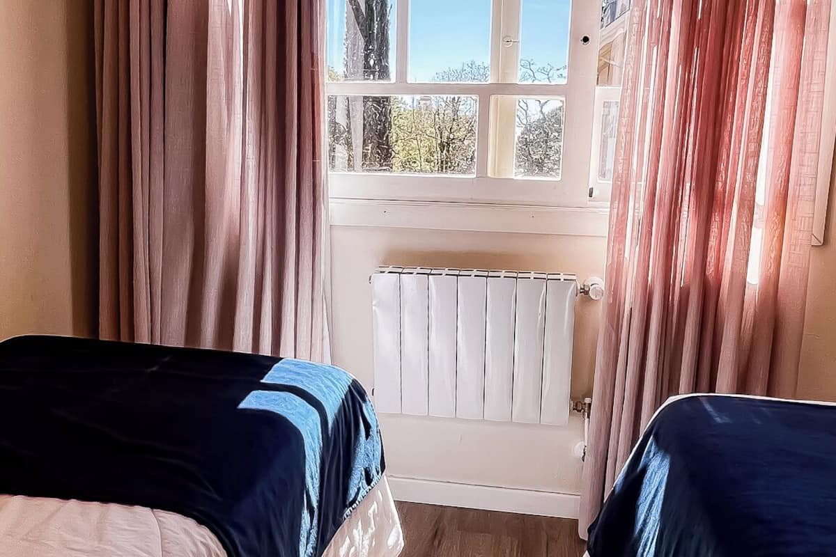 A room with two beds, a window with curtains, a radiator, and a view of trees and buildings.