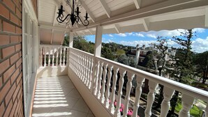 Een overdekt balkon met een witte balustrade, een zwarte kroonluchter en uitzicht op omliggende gebouwen en groen.
