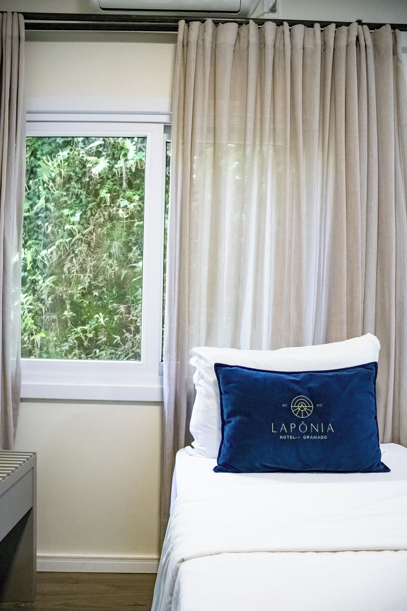 Una habitación de hotel con cama, ventana con cortinas y almohada con el nombre del hotel.
