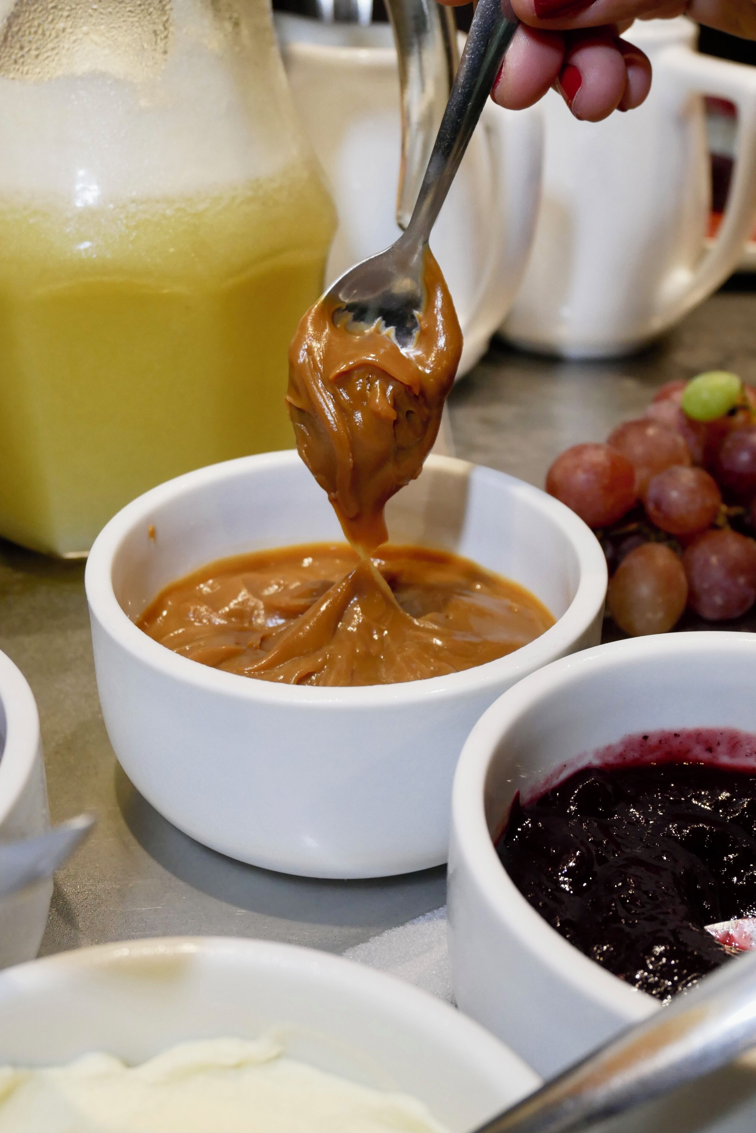 A spoonful of peanut butter being lifted from a white bowl, with a pitcher of orange juice and a bowl of red fruit jam in the background.