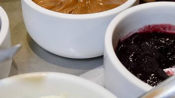 A spoonful of peanut butter being lifted from a white bowl, with a pitcher of orange juice and a bowl of red fruit jam in the background.