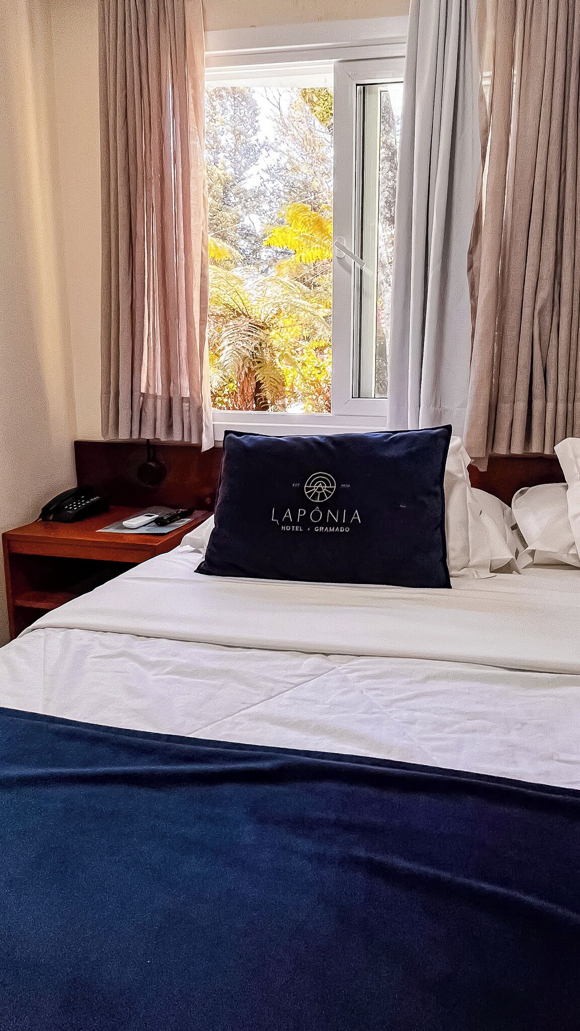 A hotel room with a bed, a nightstand with a phone, and a window with curtains.