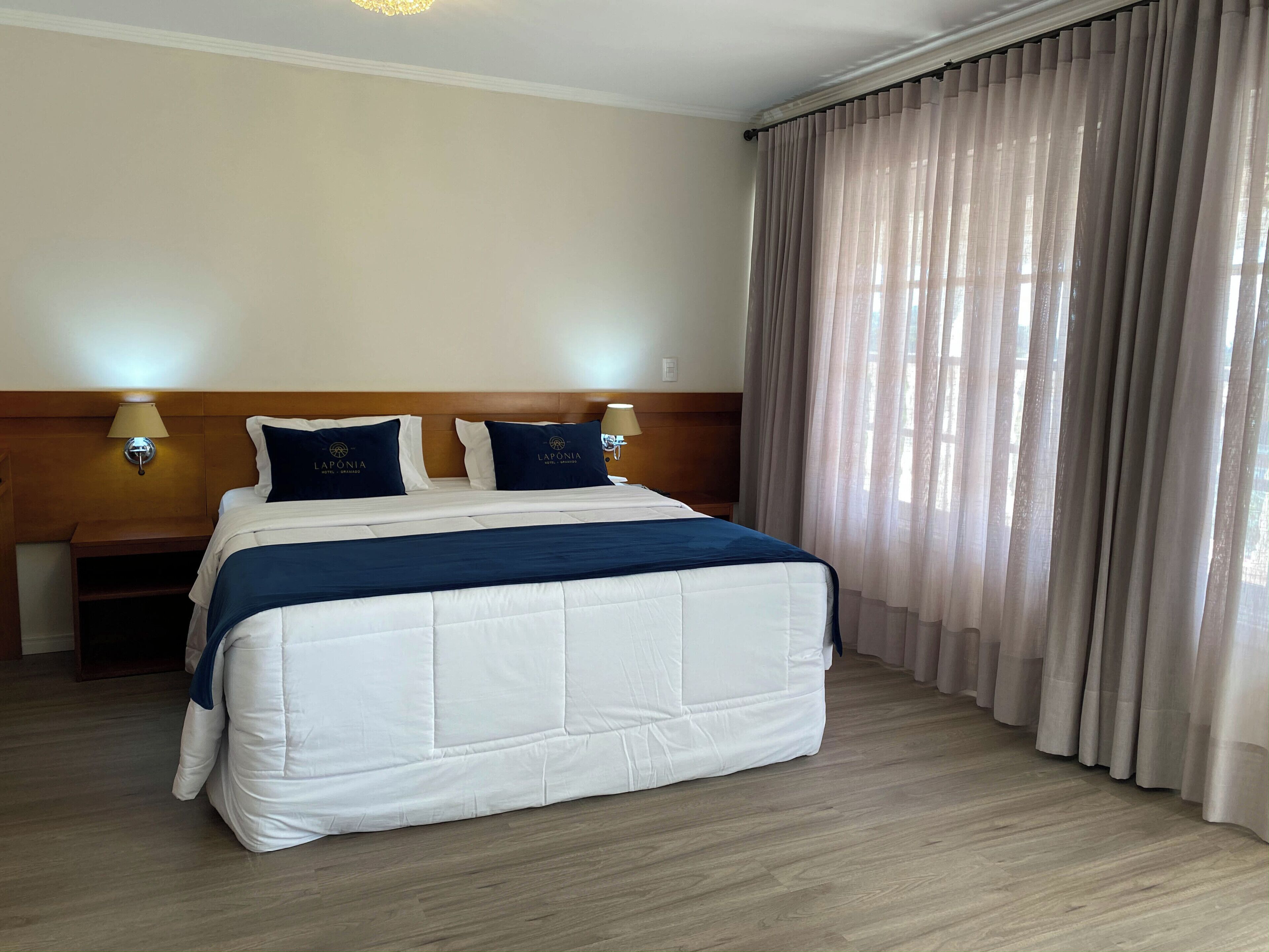 A hotel room with a large bed, two bedside lamps, a wooden headboard, and sheer curtains.