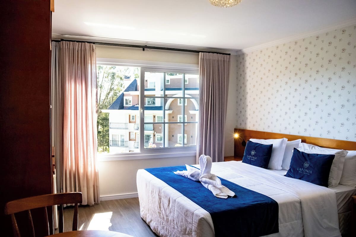 A hotel room with a large window, a bed with blue bedding, and a view of buildings outside.
