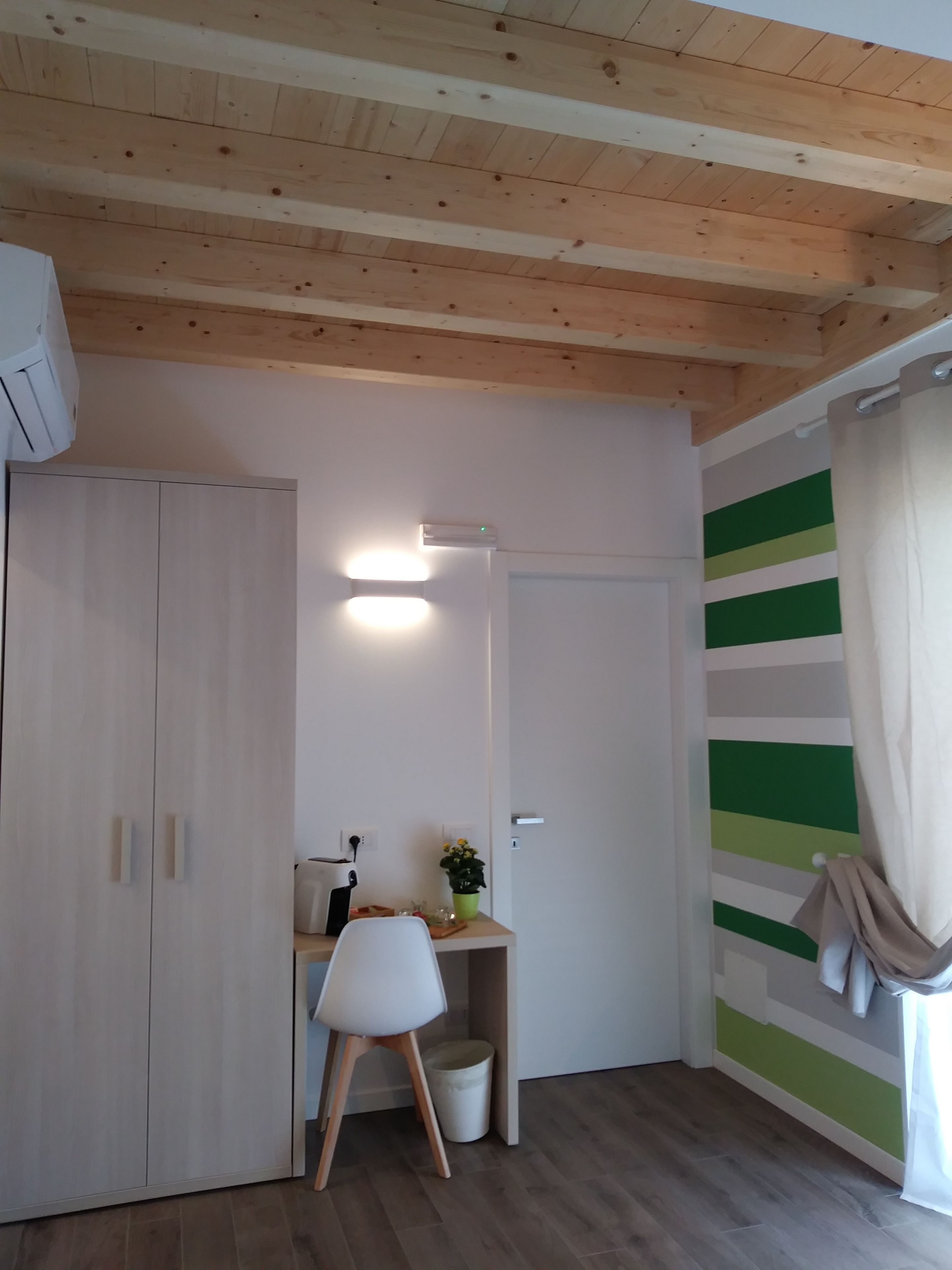 Una stanza con soffitto in legno, un'armadio bianco, una scrivania con una sedia, un cestino e una parete a righe verdi.