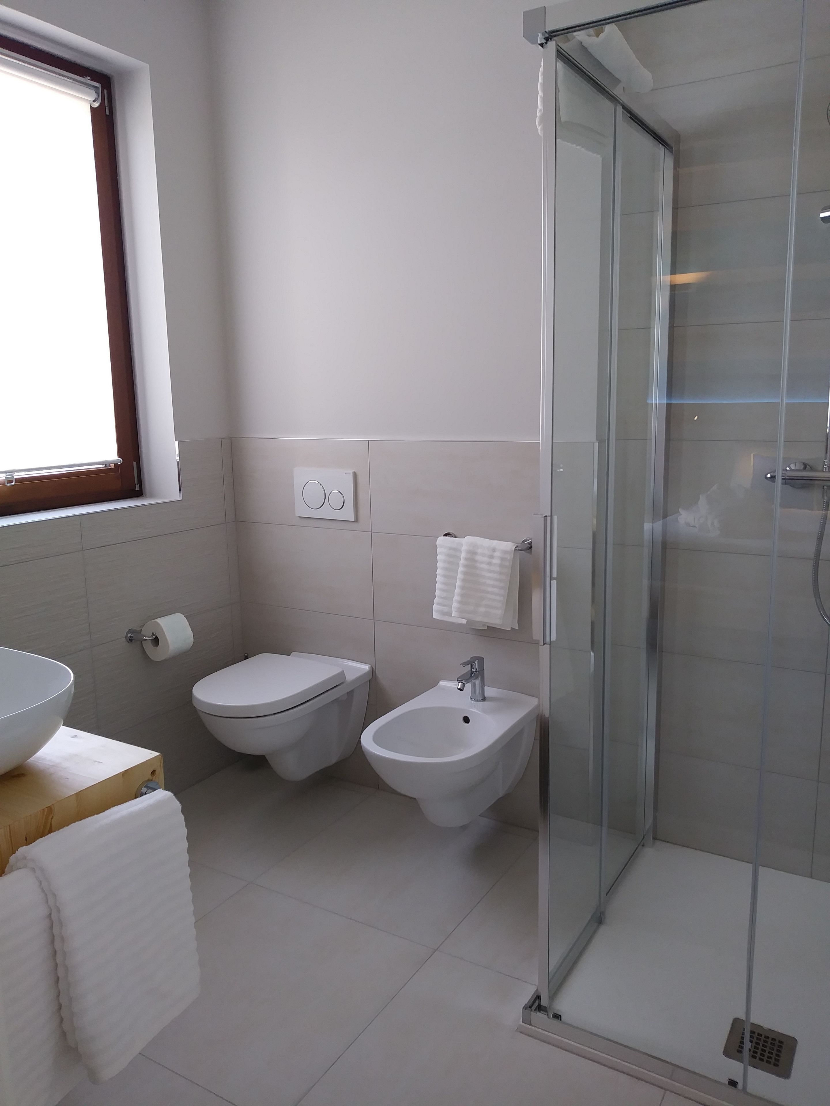 Un baño moderno con ducha de vidrio, un lavabo, dos inodoros y una ventana con persianas.