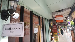 Ein Hostel namens 'ONE BANGKOK' mit einem Schild, einer Straßenszene und einem 'KERRY EXPRESS' Bushaltestellenschild.