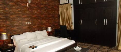 A hotel room with a large bed, a wardrobe, a nightstand with a lamp, and a small table.