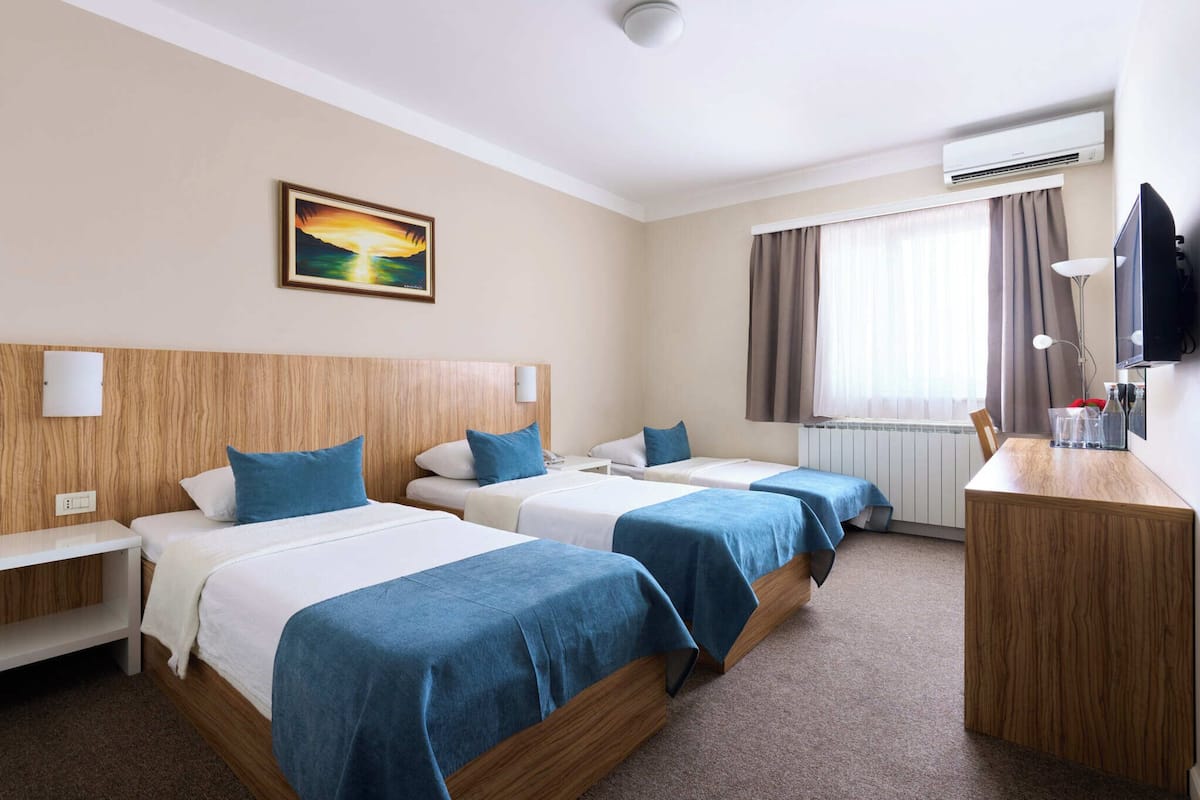 A hotel room with three beds, a desk, a TV, and a painting on the wall.