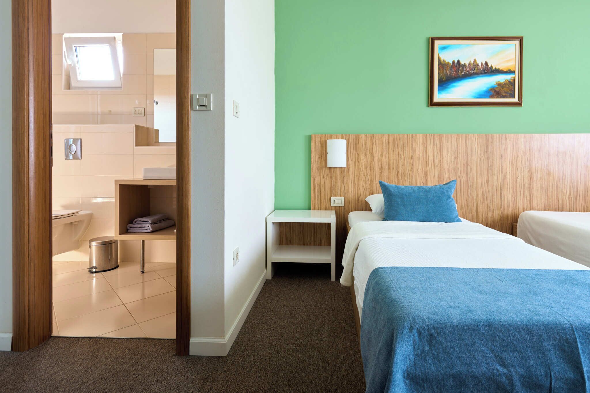 Quarto de hotel com cama, mesa de cabeceira, pintura na parede, janela e banheiro visível por uma porta aberta.